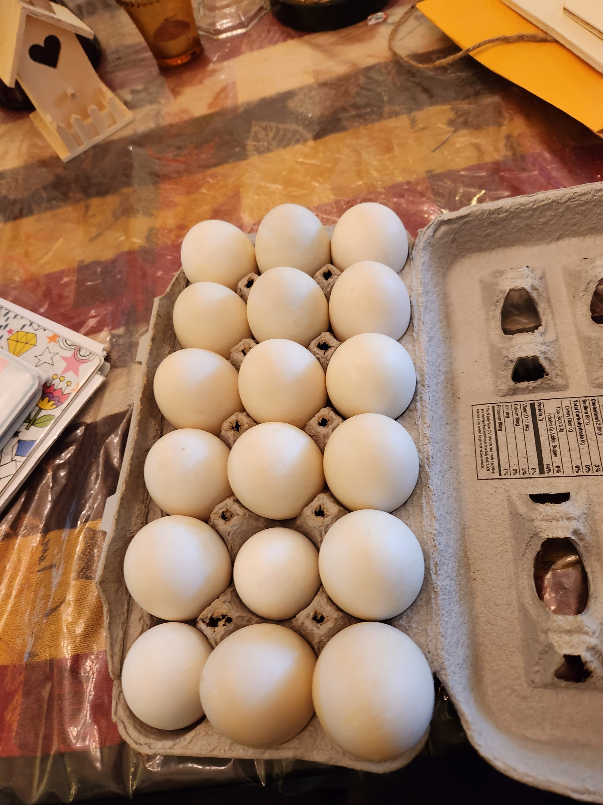 A carton of white eggs placed on a table with a colorful tablecloth. Nearby are some papers and a small decorative white house with a heart cutout.