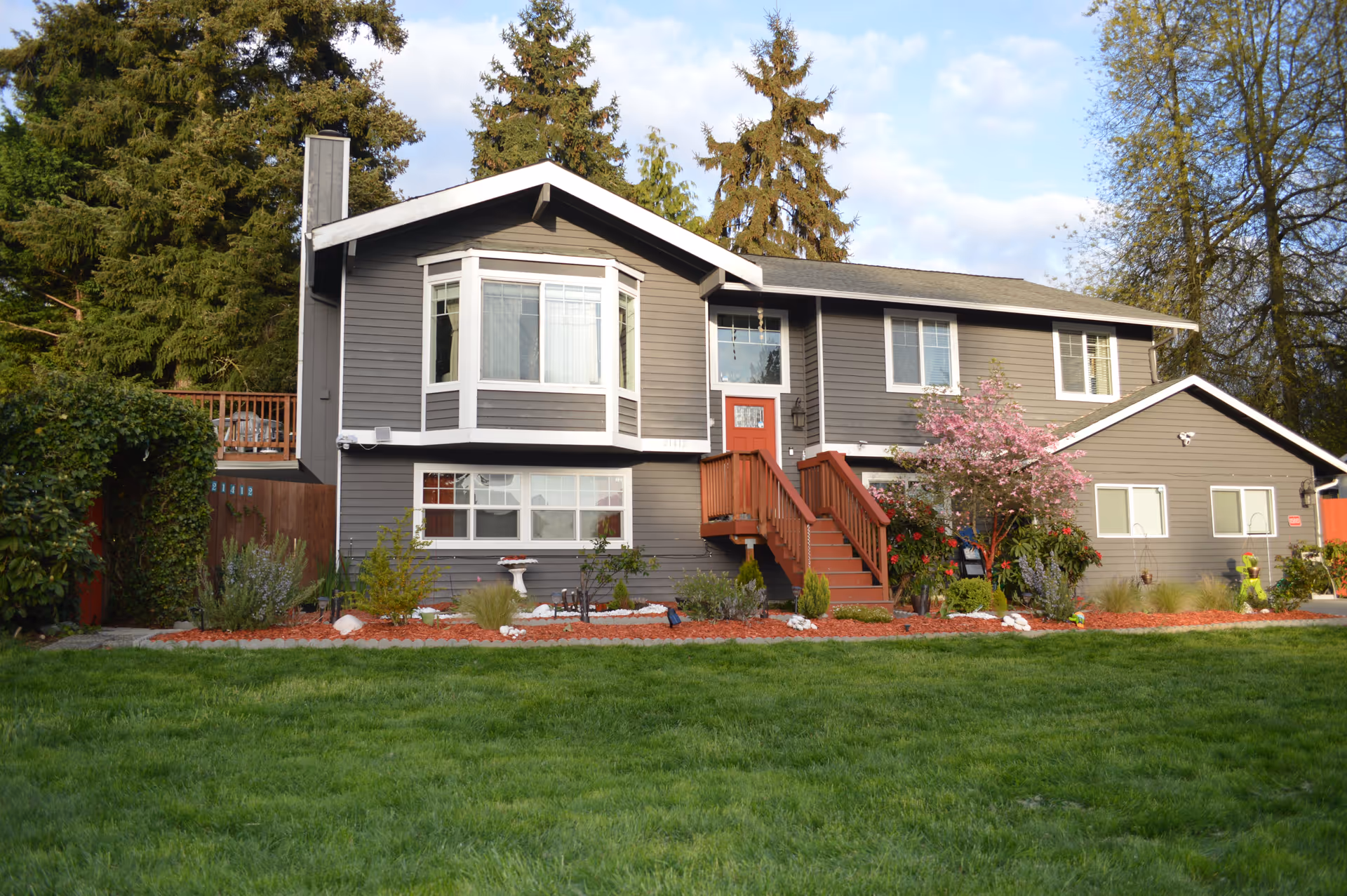 Exterior view of a two-story gray house with white trim and a red front door. The house has a well-maintained garden with various plants, flowers, and a small tree with pink blossoms. There is a green lawn in the foreground and tall evergreen trees in the background under a partly cloudy sky.