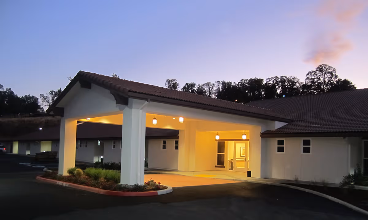 Exterior view of the entrance to Sonora Convalescent Hospital at dusk, showing a covered drop-off area with lights on and a tiled roof, surrounded by trees and a parking area.