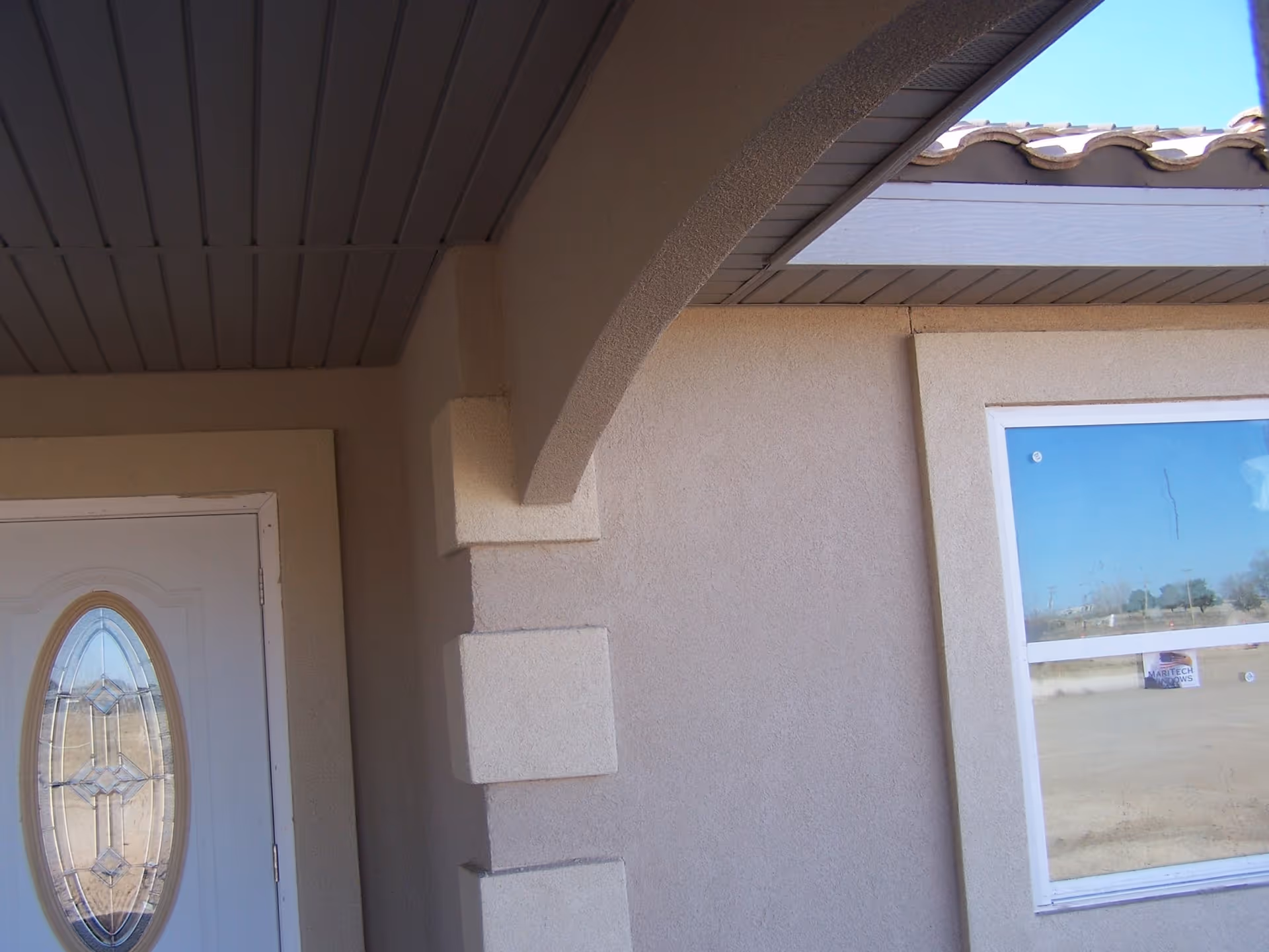 Close-up view of the exterior entrance of a building showing a white door with an oval decorative glass panel, a beige stucco wall with architectural details, a window reflecting the outdoor landscape, and part of a tiled roof under a blue sky.