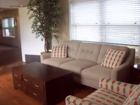 Cozy senior living lounge with beige sofa, patterned chairs, a coffee table, and a plant.
