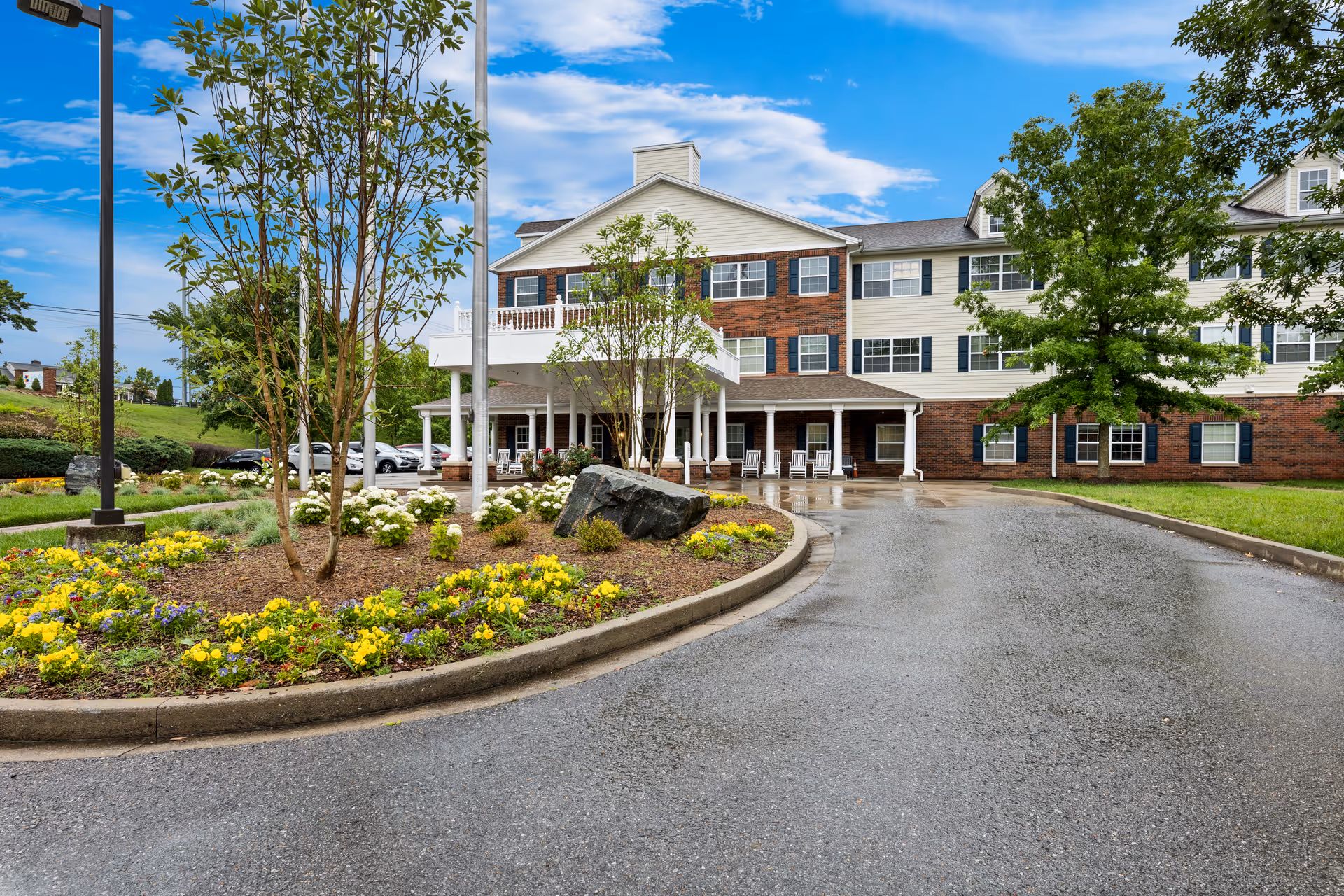 Front exterior view of Vitality Living Hendersonville, showing a three-story building with a combination of brick and light-colored siding. There is a covered entrance with white columns and rocking chairs, a curved driveway, landscaped flower beds with yellow and white flowers, and trees under a partly cloudy blue sky.