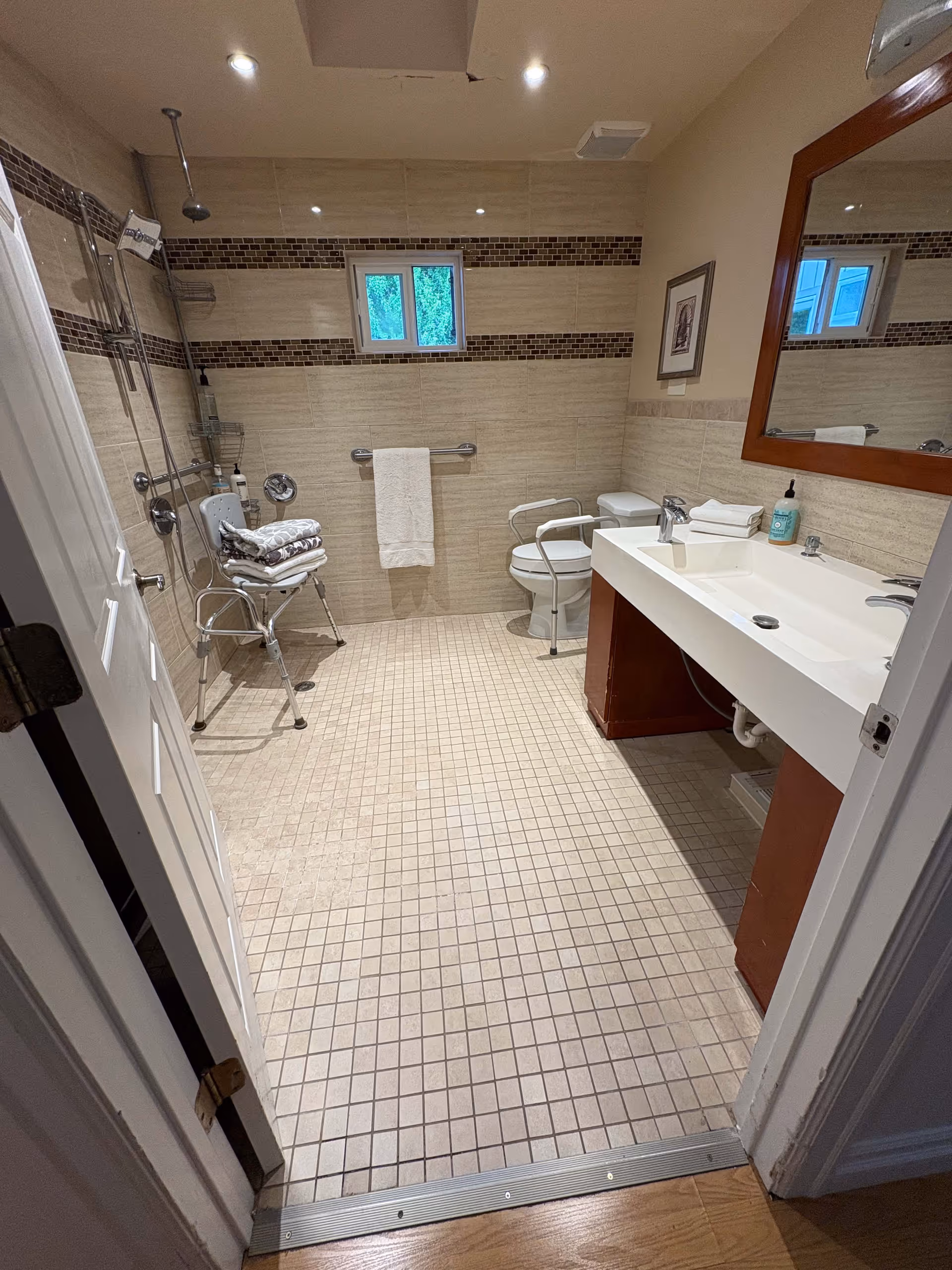 A spacious bathroom with beige tiled floor and walls, featuring a walk-in shower with a shower chair and grab bars, a toilet with support rails, a large white sink with a wooden base, a mirror above the sink, a towel hanging on a rail, and a small window providing natural light.