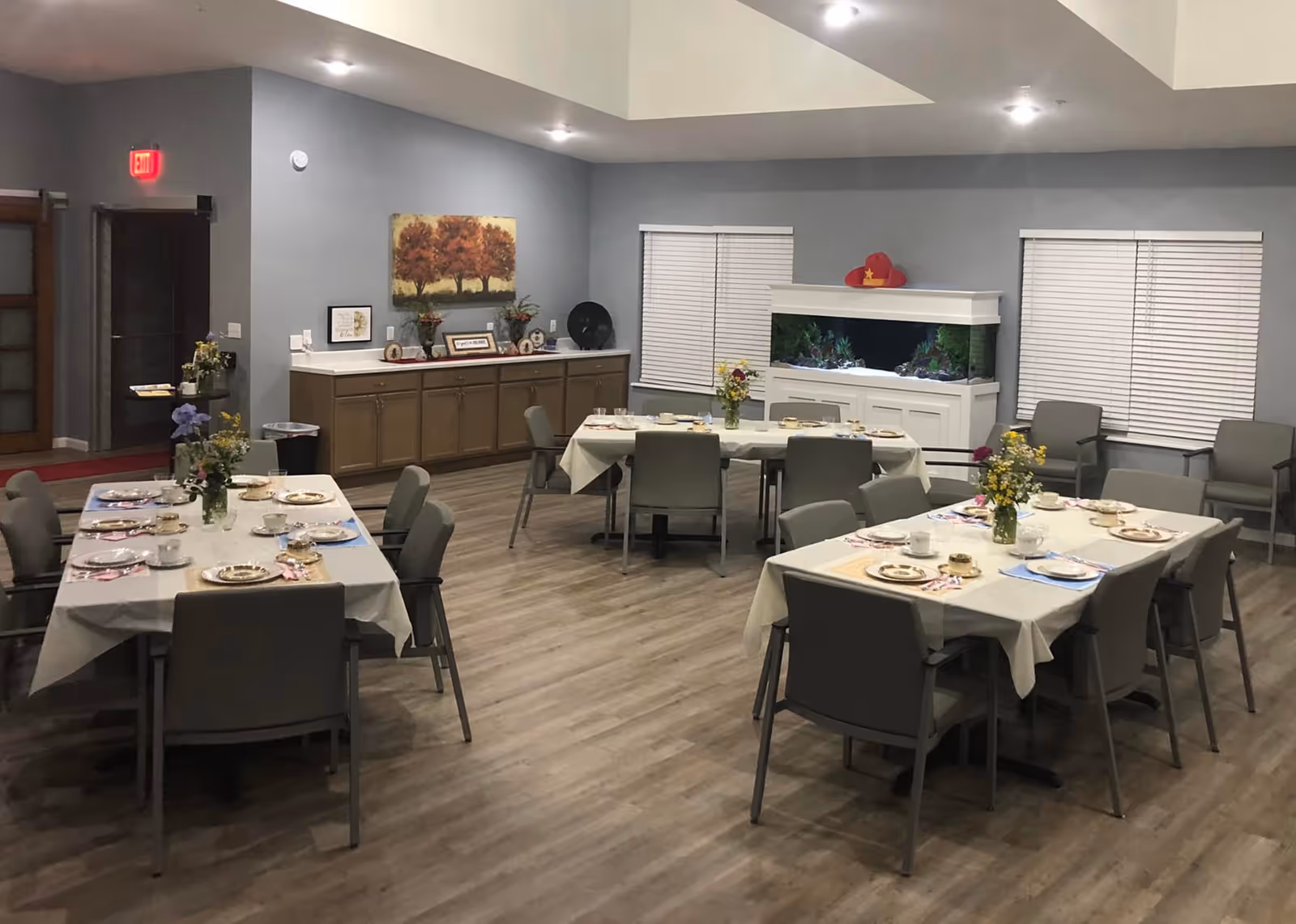 A dining room with three tables covered with white tablecloths, each set with plates, cups, and utensils. There are gray chairs around the tables and flower arrangements as centerpieces. The room has wood flooring, gray walls, two windows with blinds, a large fish tank on a white cabinet, and a sideboard with decorative items and a painting of trees on the wall.