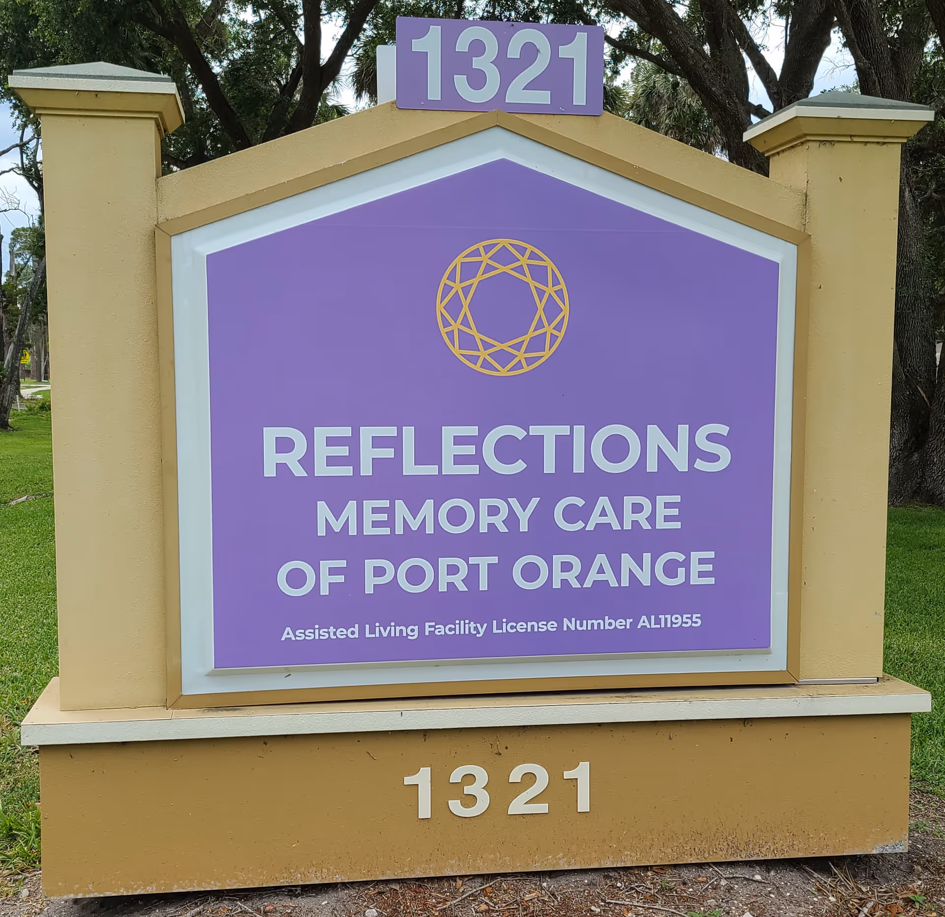 Outdoor sign for Reflections Memory Care of Port Orange with the address number 1321 displayed at the top and bottom. The sign is purple with white text and a gold geometric logo above the facility name. The sign is mounted on a beige structure with trees and grass in the background.