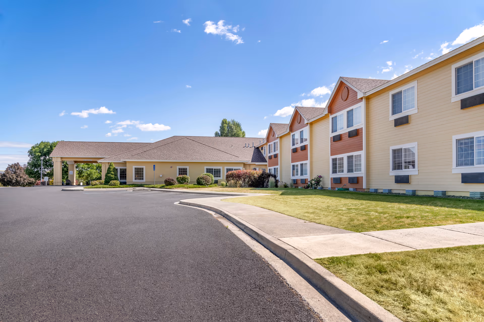 Exterior view of a senior living facility building with a paved driveway, green lawn, and clear blue sky with some clouds.