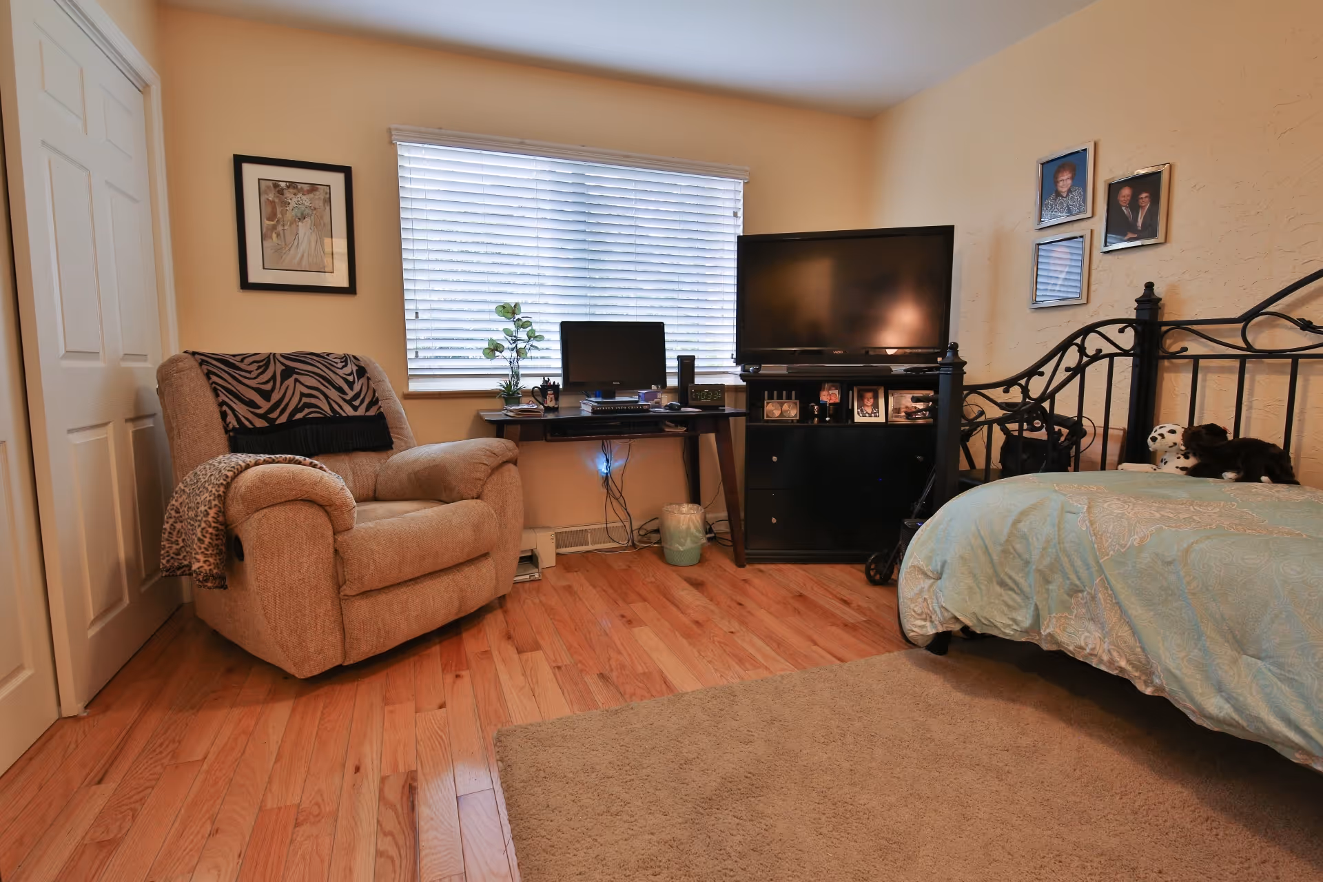 A cozy bedroom featuring a beige recliner chair with a zebra-patterned throw, a black metal bed frame with light blue bedding, a black TV stand with a flat-screen television, and a small desk with a computer monitor and keyboard in front of a window with white blinds. The room has wooden flooring, a beige rug, and framed photos and artwork on the walls.