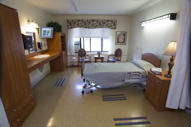 A clean and well-lit senior living bedroom with a hospital-style adjustable bed covered with a light gray blanket. The room features wooden furniture including a nightstand with a lamp and telephone, a long counter with a sink, a mounted TV, and a paper towel dispenser. There are two chairs and a small round table near a window with floral valance and white curtains. The floor has beige linoleum with black stripes, and the walls are light-colored with a framed picture hanging beside the window.