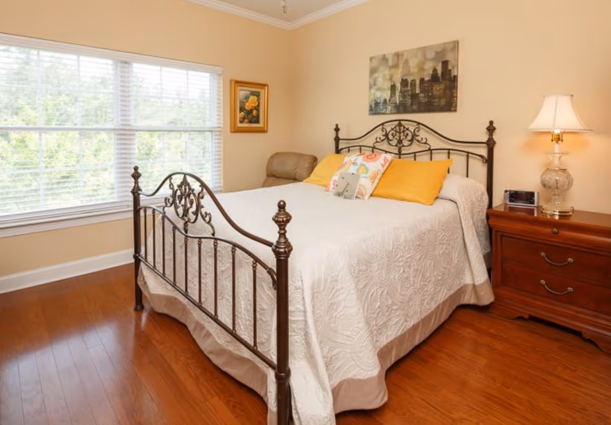 Neatly made bedroom with an ornate metal bed frame, bedside table and lamp beside a large window.