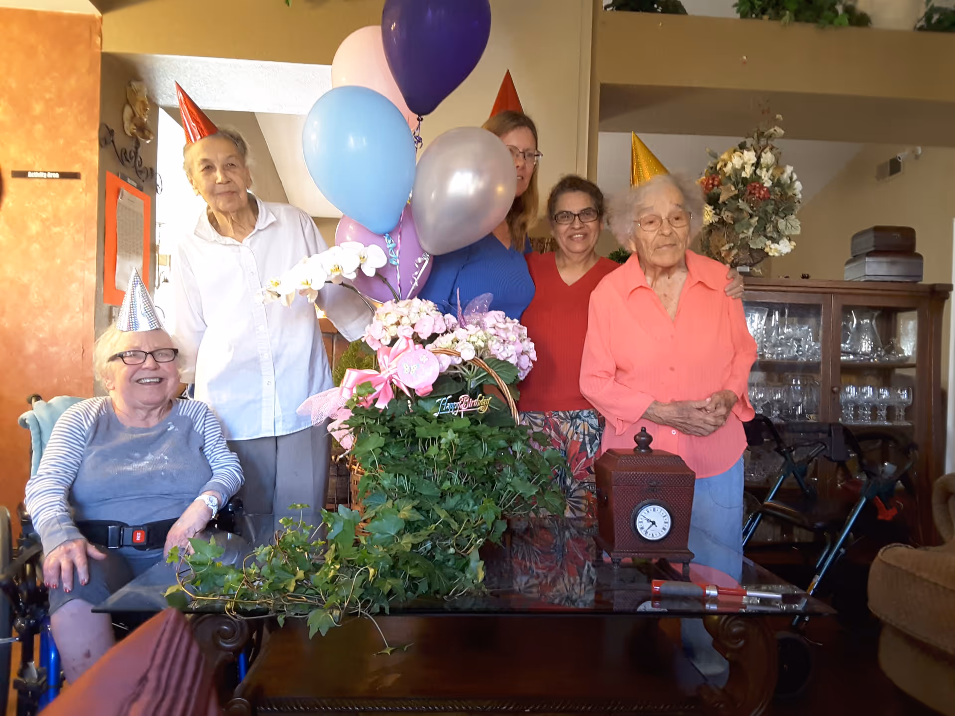 A group of five elderly women and one middle-aged woman celebrating a birthday indoors. They are wearing colorful party hats and standing or sitting around a table with a bouquet of flowers, balloons, and a small clock. The room has warm lighting and a cabinet with glassware in the background.