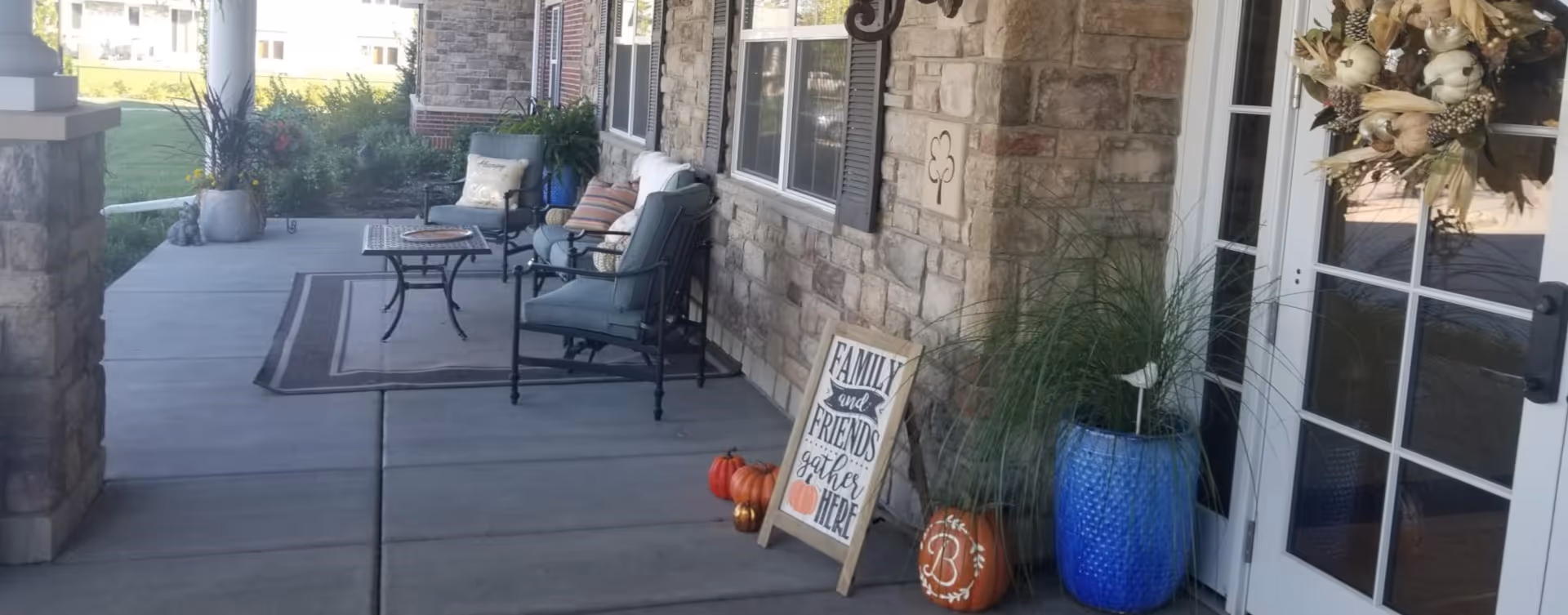 Outdoor covered patio area with stone walls and concrete floor. There are cushioned chairs and a small table on a rug, potted plants in blue pots, decorative pumpkins, and a sign that reads 'Family and Friends gather here'. A wreath hangs on the glass door.