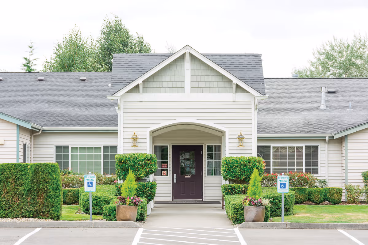 Front exterior view of South Pointe Assisted Living facility showing a single-story building with a gray shingled roof, white siding, a central entrance with a dark door, flanked by windows and decorative shrubs. There are two reserved handicap parking signs in front of the building.