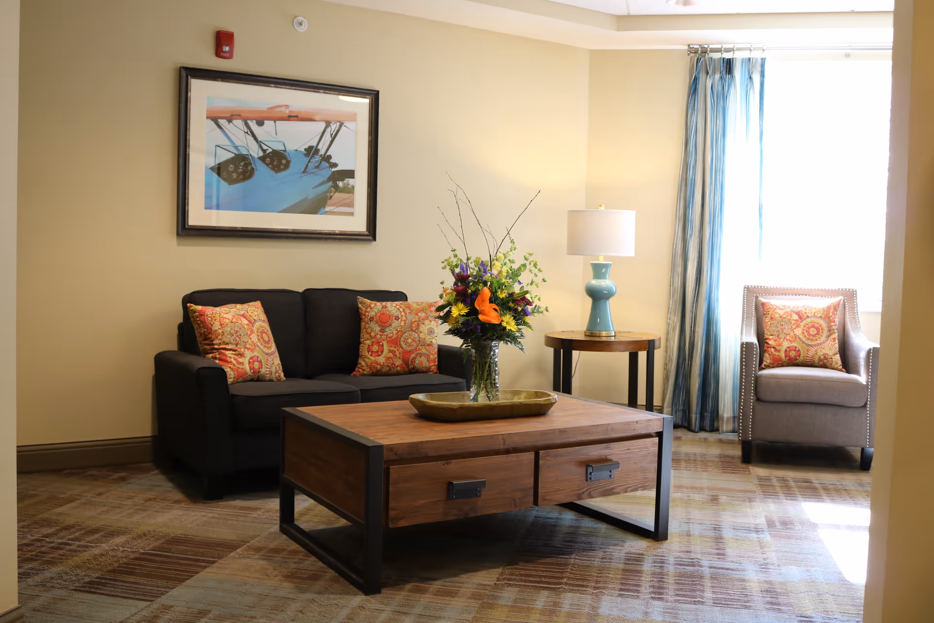 A cozy living room area with a dark gray sofa adorned with three patterned cushions, a wooden coffee table with a decorative tray and a vase of colorful flowers, a round side table with a blue lamp, and a gray armchair with a patterned cushion near a window with blue curtains.