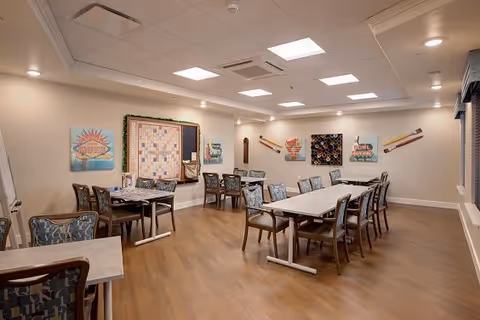 Well-lit communal dining room with multiple tables and chairs, wall artwork and wooden floors.
