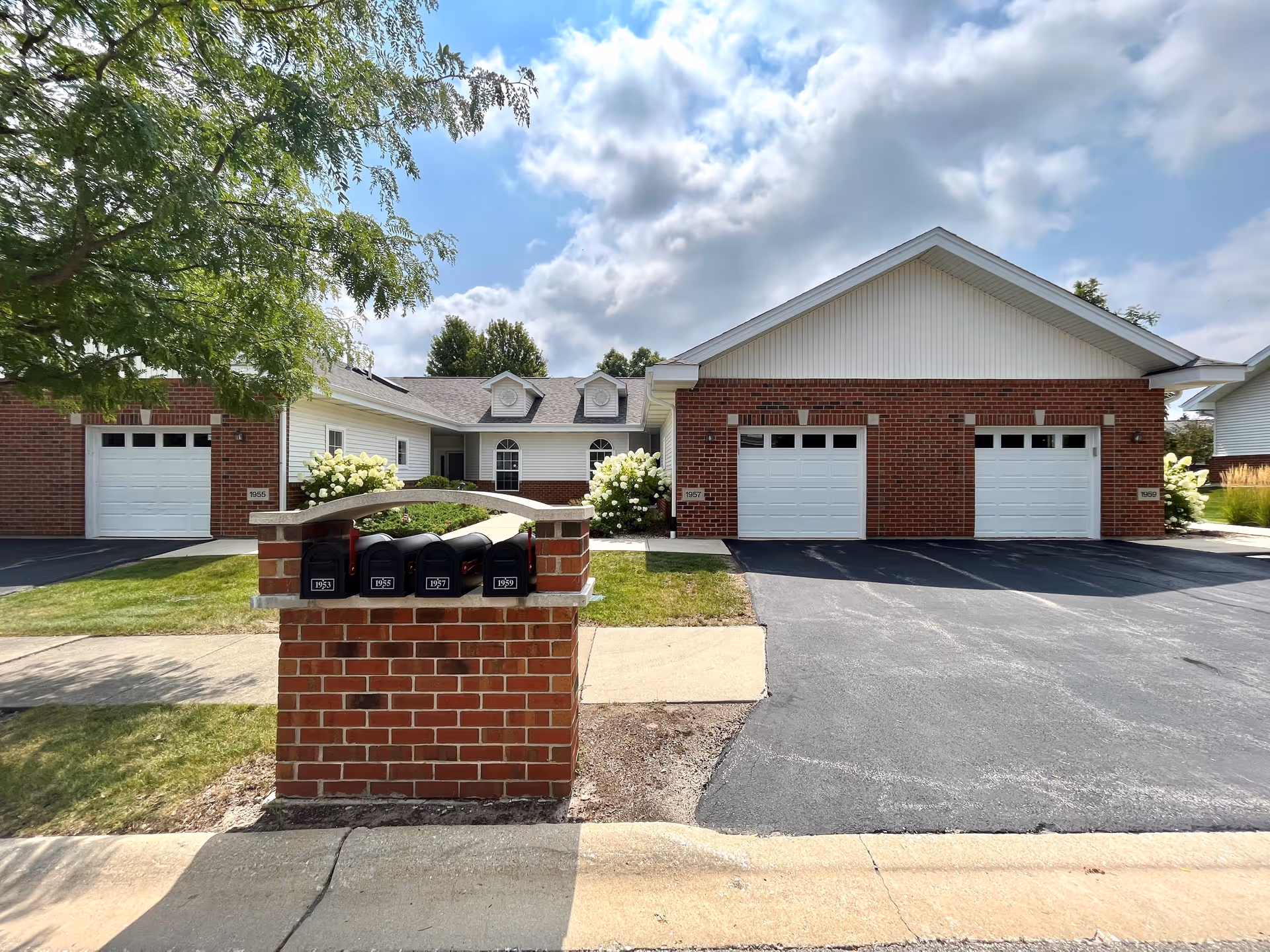 Exterior view of a residential building with brick and white siding, featuring three white garage doors and a brick mailbox structure with four black mailboxes numbered 1931, 1955, 1957, and 1959. There is a tree on the left side and a partly cloudy sky above.