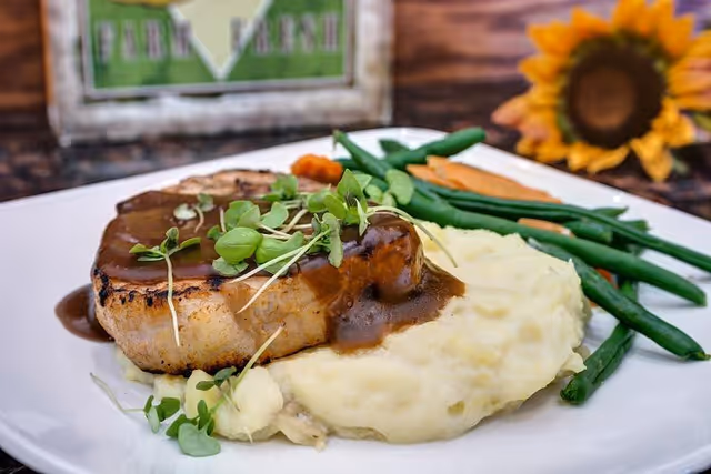 A plated meal featuring a grilled pork chop topped with brown gravy and microgreens, served with mashed potatoes and steamed green beans and carrots on a white plate. A sunflower and a decorative sign are visible in the background.