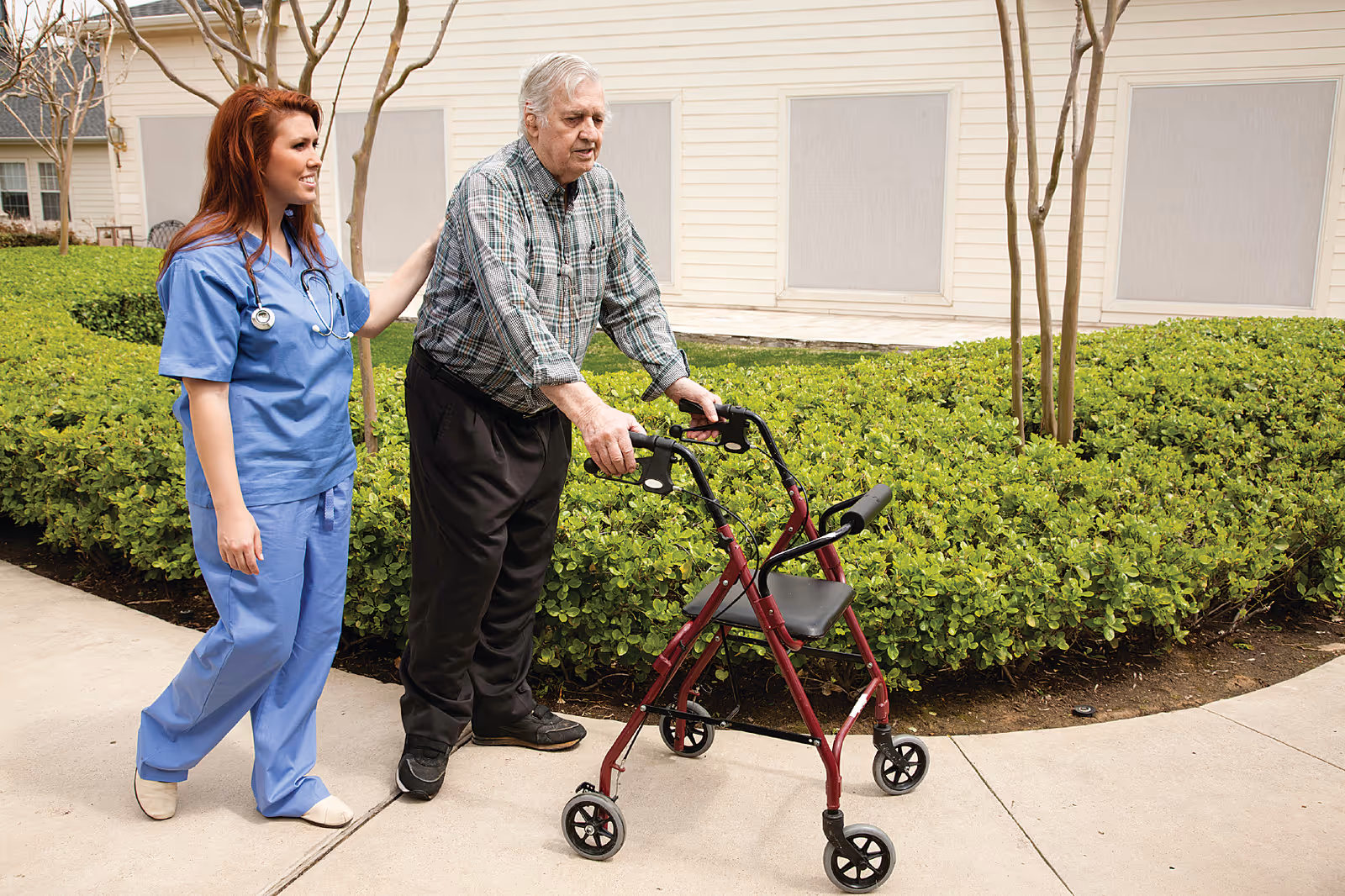 A female healthcare worker in blue scrubs assists an elderly man using a red walker as they walk on a paved path outside near green bushes and a beige building.
