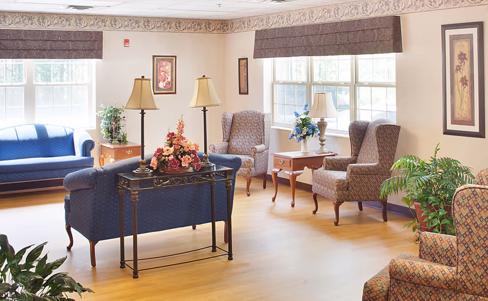 A bright and cozy living room in a senior living facility with large windows letting in natural light. The room features blue upholstered sofas, patterned armchairs, wooden side tables with lamps, floral arrangements, framed artwork on the walls, and potted plants on the floor.