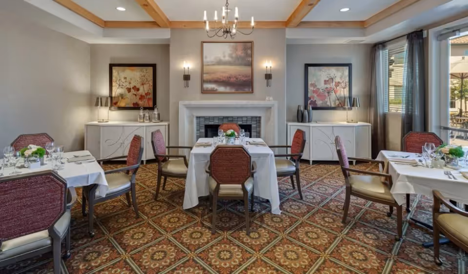 A neatly set dining room with tables and chairs arranged around a central fireplace, chandelier, and framed artwork.