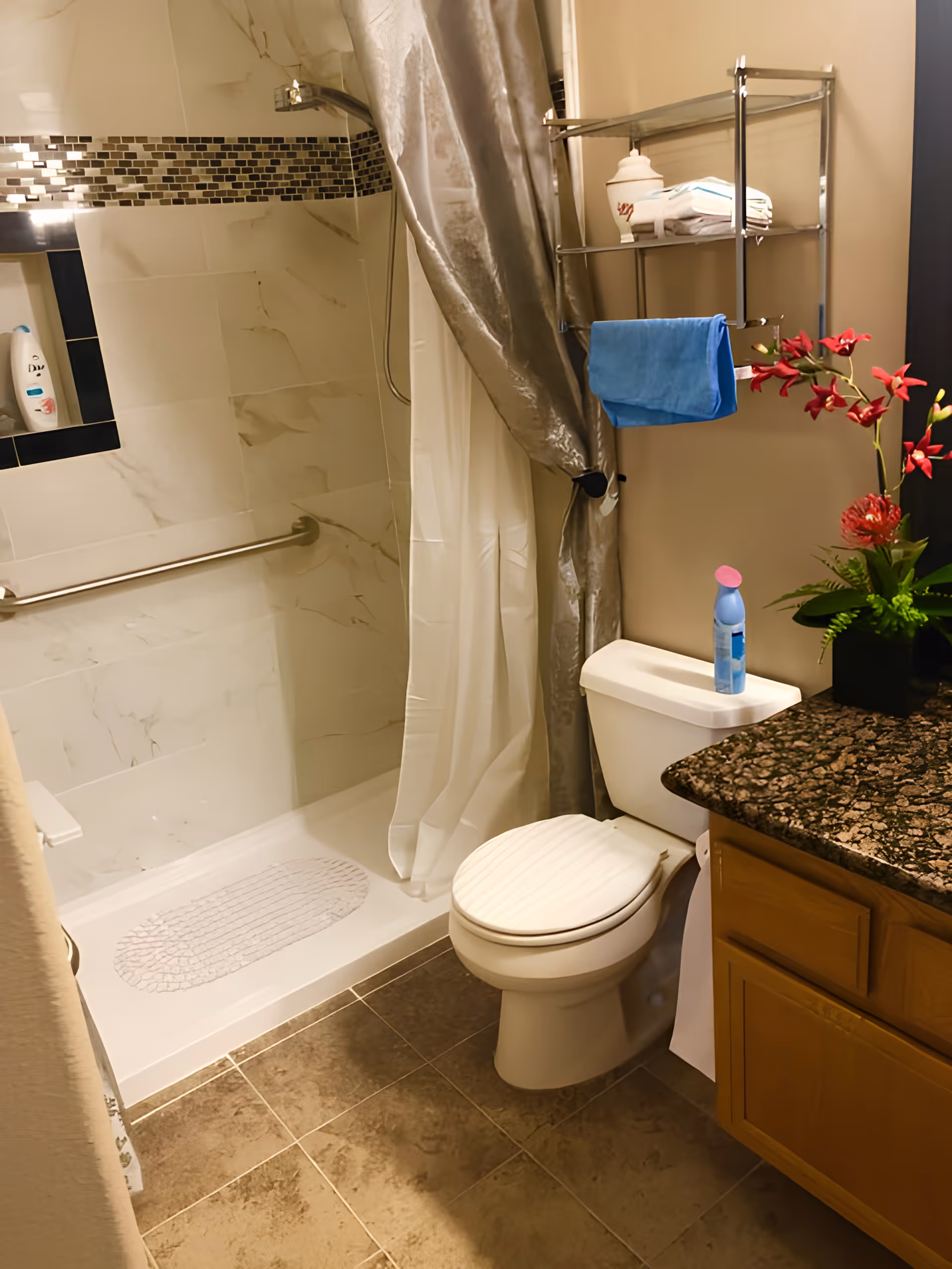 A clean bathroom featuring a walk-in shower with a grab bar and a white textured mat, a toilet with a blue spray bottle on the tank, a metal shelf holding folded towels and a blue hand towel, a granite countertop with a wooden cabinet below, and a decorative plant with red flowers.