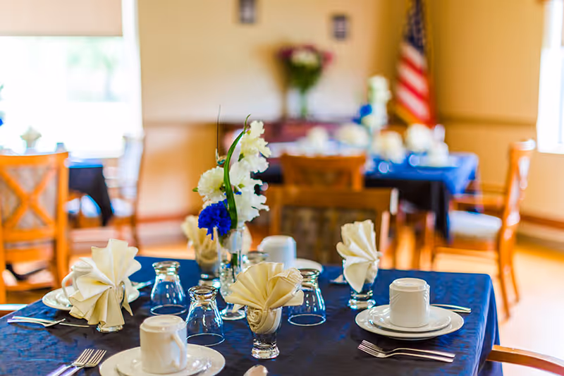 A dining room table with a blue tablecloth set with folded napkins, plates, cups, glassware, and a vase of flowers.