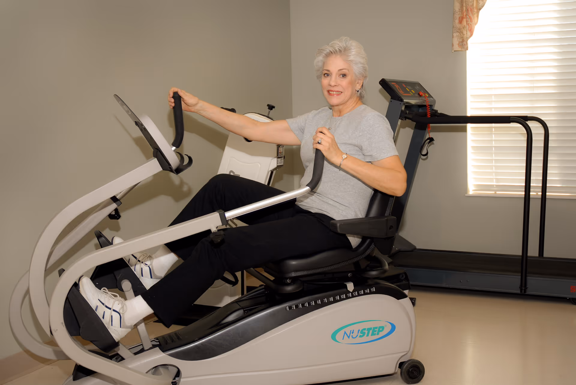 An elderly woman with short white hair wearing a gray t-shirt and black pants is exercising on a NuStep recumbent cross trainer machine in a room with beige walls and a window with blinds. A treadmill is visible in the background.