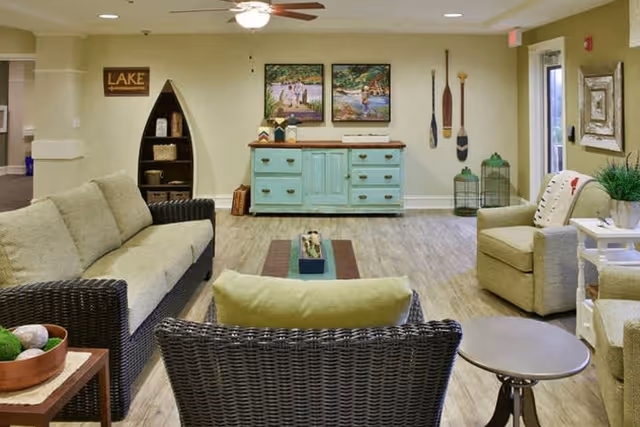 A cozy living room area in a senior living facility featuring wicker furniture with beige cushions, a light blue wooden cabinet against the wall, decorative paddles hanging on the wall, two framed paintings, and a ceiling fan above. The room has light-colored walls and wood flooring, with a small round table and some plants adding to the decor.