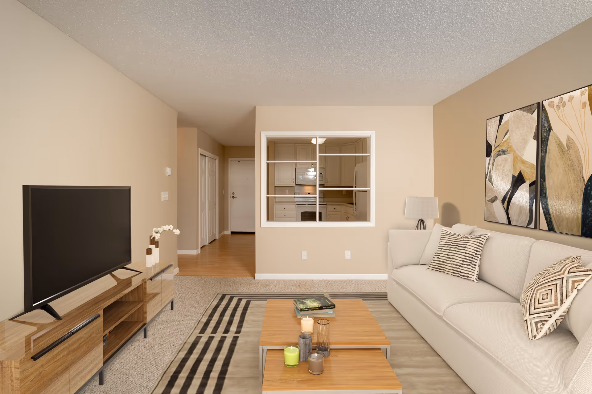 A cozy living room with a beige sofa adorned with patterned pillows, a wooden coffee table with candles and books, a flat-screen TV on a wooden stand, and abstract artwork on the wall. A window opening in the wall reveals a kitchen area with white cabinets and appliances. The room has beige walls and carpeted flooring, with a hallway leading to a white door in the background.