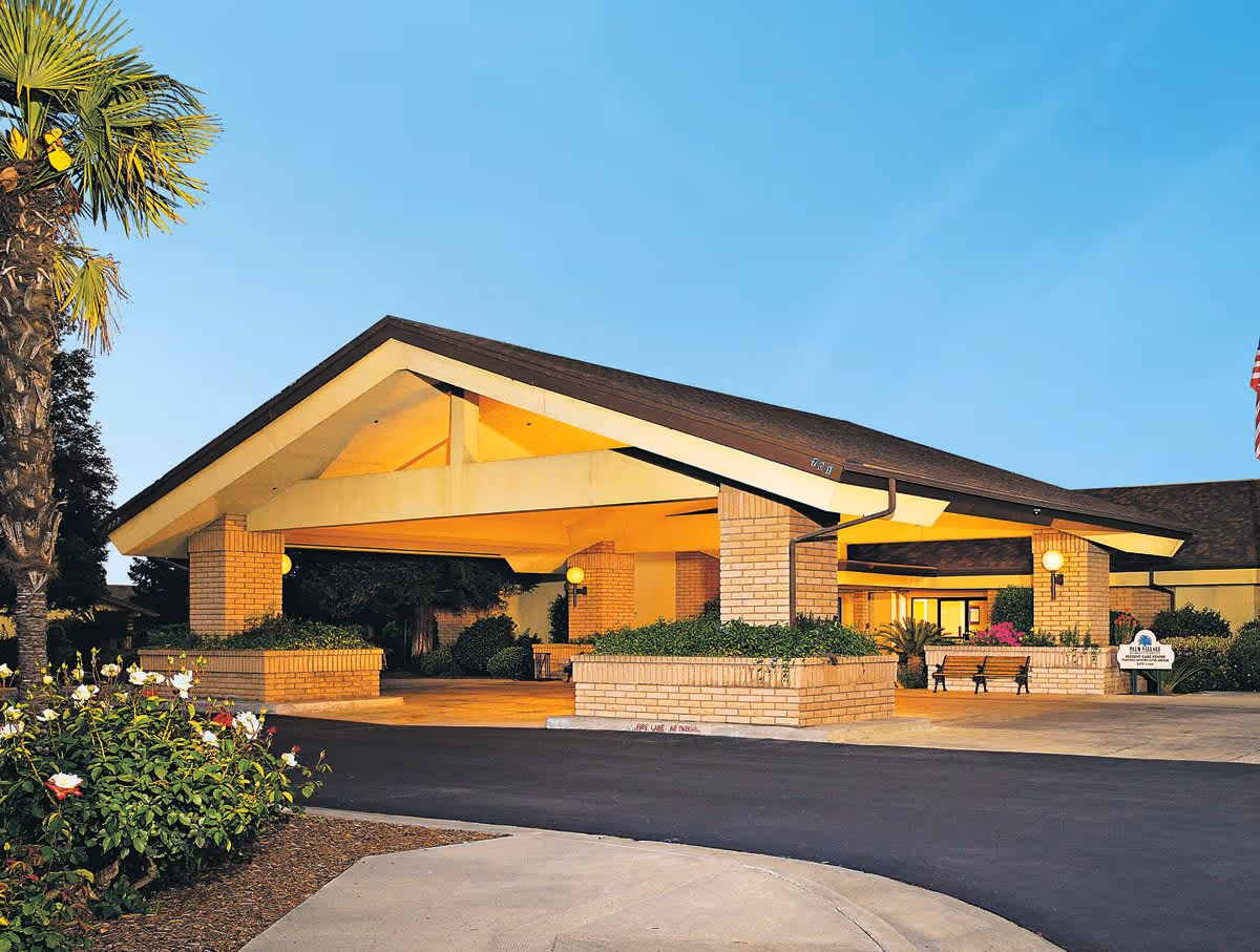 Front entrance of Palm Village Retirement Community with a covered porte-cochère, palm tree, and landscaped beds.