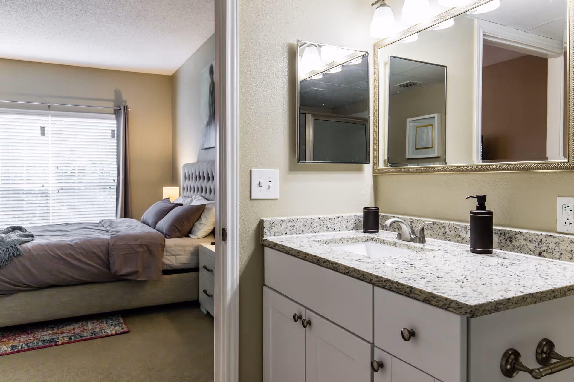 View of a bedroom and bathroom area in a senior living facility. The bedroom features a bed with gray bedding, pillows, a nightstand with a lamp, and a window with blinds and curtains. The bathroom area has a granite countertop with a sink, a soap dispenser, a toothbrush holder, a large mirror, and overhead lighting.