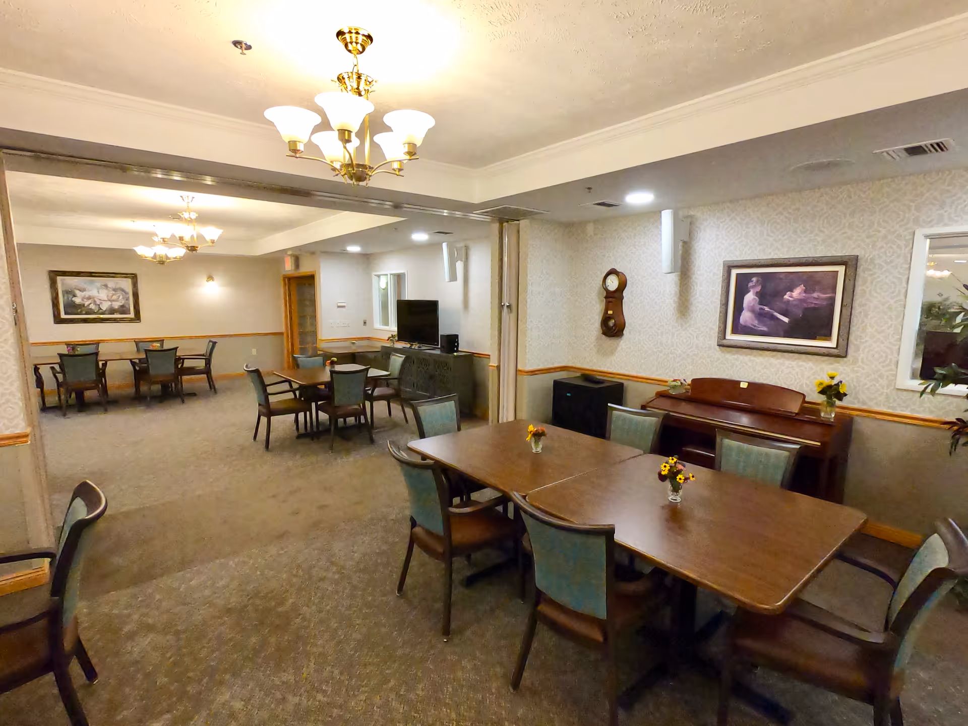 Interior view of a senior living facility common area with multiple tables and chairs arranged for seating. The room features carpeted floors, wallpapered walls with framed artwork, a wall clock, a piano, and ceiling light fixtures. The space appears to be designed for dining or social activities.