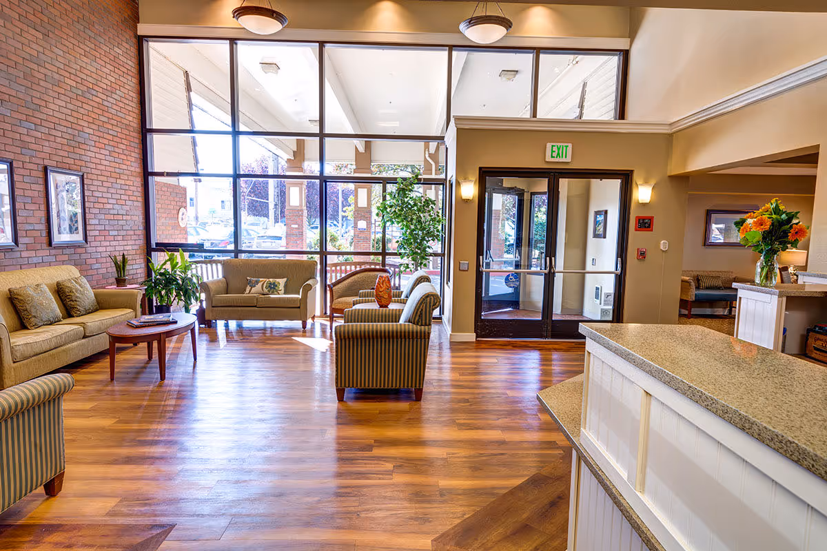 A bright and spacious senior living facility lobby with wooden flooring, beige sofas, striped armchairs, a coffee table, potted plants, and large windows letting in natural light. There is a reception desk on the right side and glass doors leading outside.