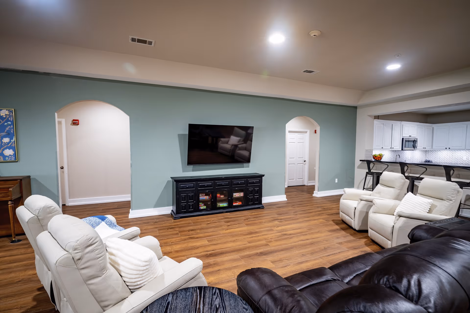 Spacious communal living room with multiple recliners, a wall-mounted TV over a console, wood floors, and an open view into a kitchen area.