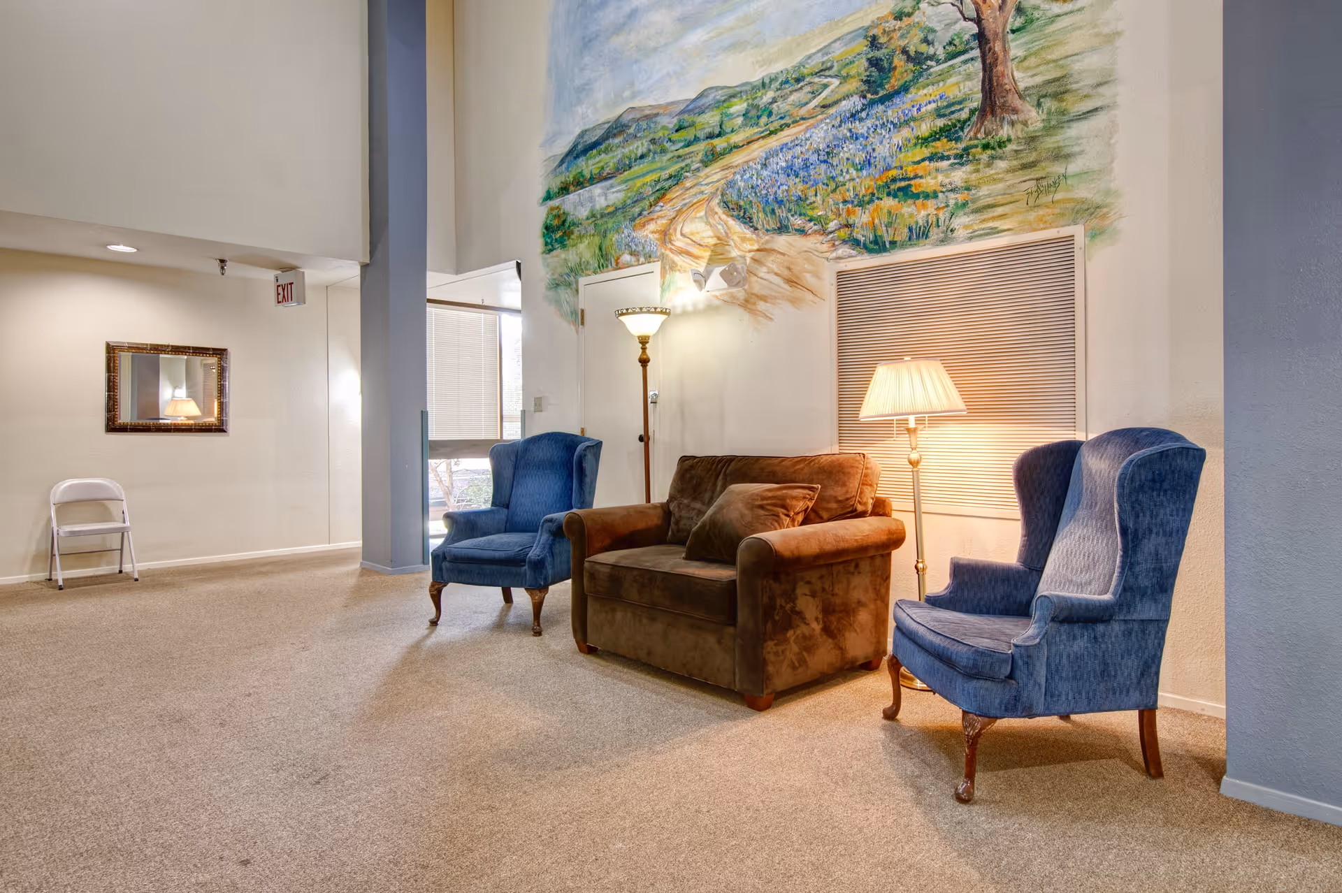 A cozy seating area in a senior living facility with two blue upholstered armchairs and a brown loveseat arranged around two floor lamps. The wall behind the seating features a large mural of a scenic landscape with a winding path, trees, and flowers. The room has beige carpeting and light-colored walls, with a mirror and a white folding chair visible in the background.