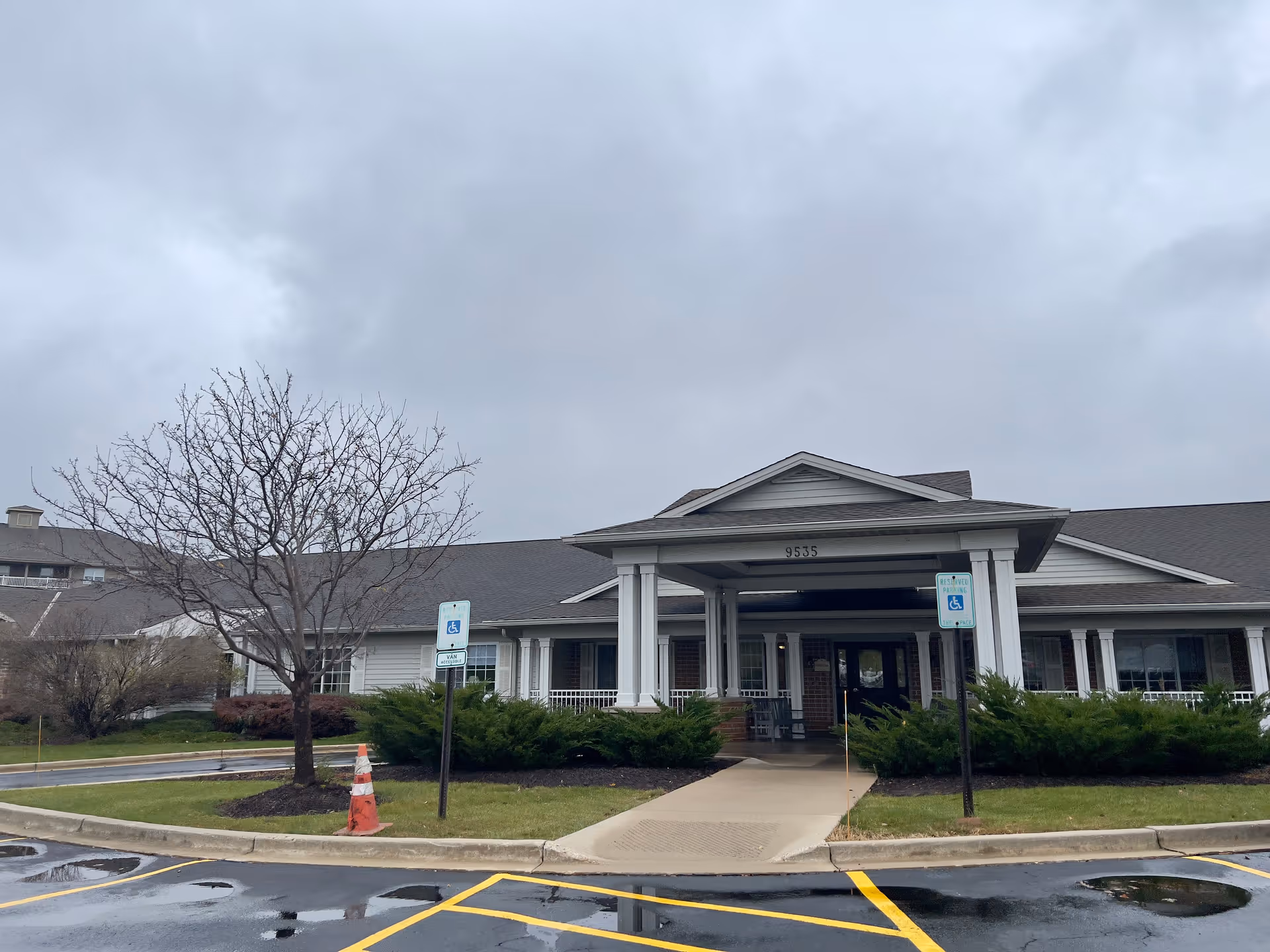 Exterior view of Brenwood Park Assisted Living facility showing the main entrance with a covered porch supported by white columns. There are two reserved handicapped parking signs in front, a leafless tree, bushes, and a wet parking lot with yellow painted lines under a cloudy sky.