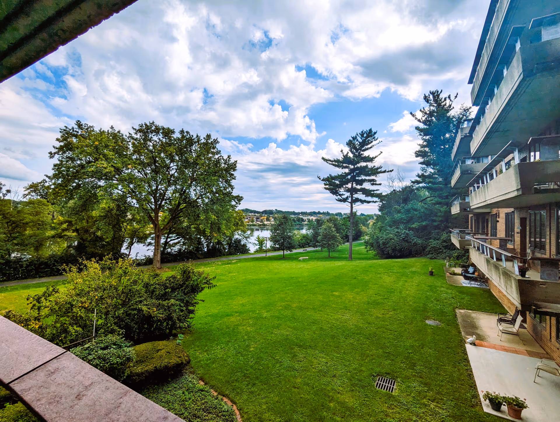 A scenic view from a balcony at Plum Landing Retirement Community, showcasing a lush green lawn, trees, and a glimpse of a lake under a partly cloudy sky.