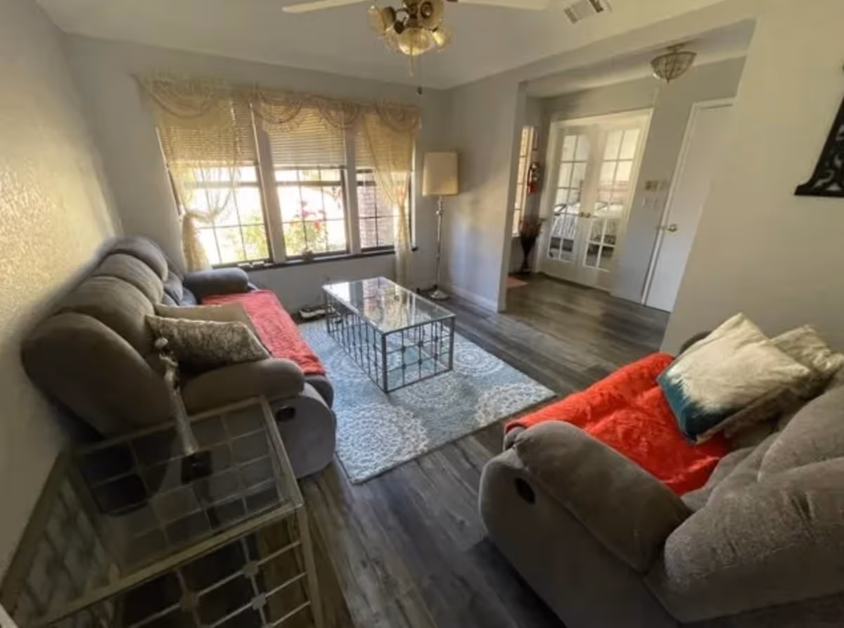 A cozy living room with two gray recliner sofas covered partially with red throws, a glass coffee table on a patterned rug, a floor lamp, and large windows with sheer curtains allowing natural light to fill the room. The room has wood flooring and a ceiling fan with lights.