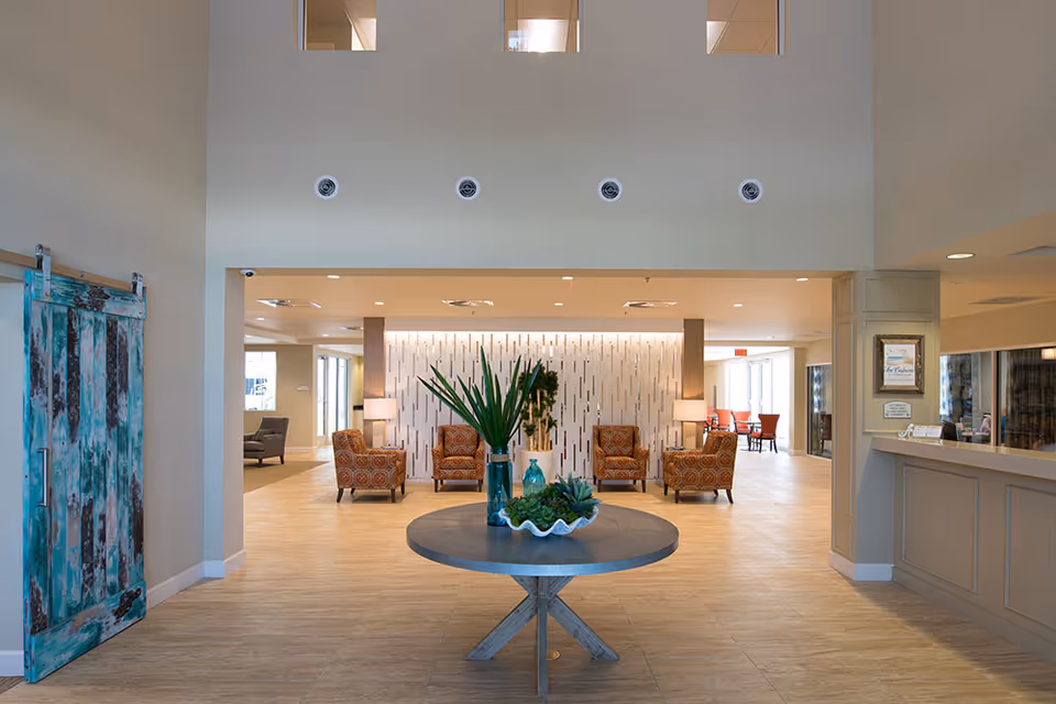 Spacious and well-lit senior living facility common area with a round table in the center decorated with plants and vases. Four patterned armchairs are arranged against a decorative wall with vertical accents. To the left, there is a rustic sliding barn door with a distressed turquoise finish. On the right, a reception desk with a framed sign and brochures is visible. The floor is tiled and the ceiling has recessed lighting.