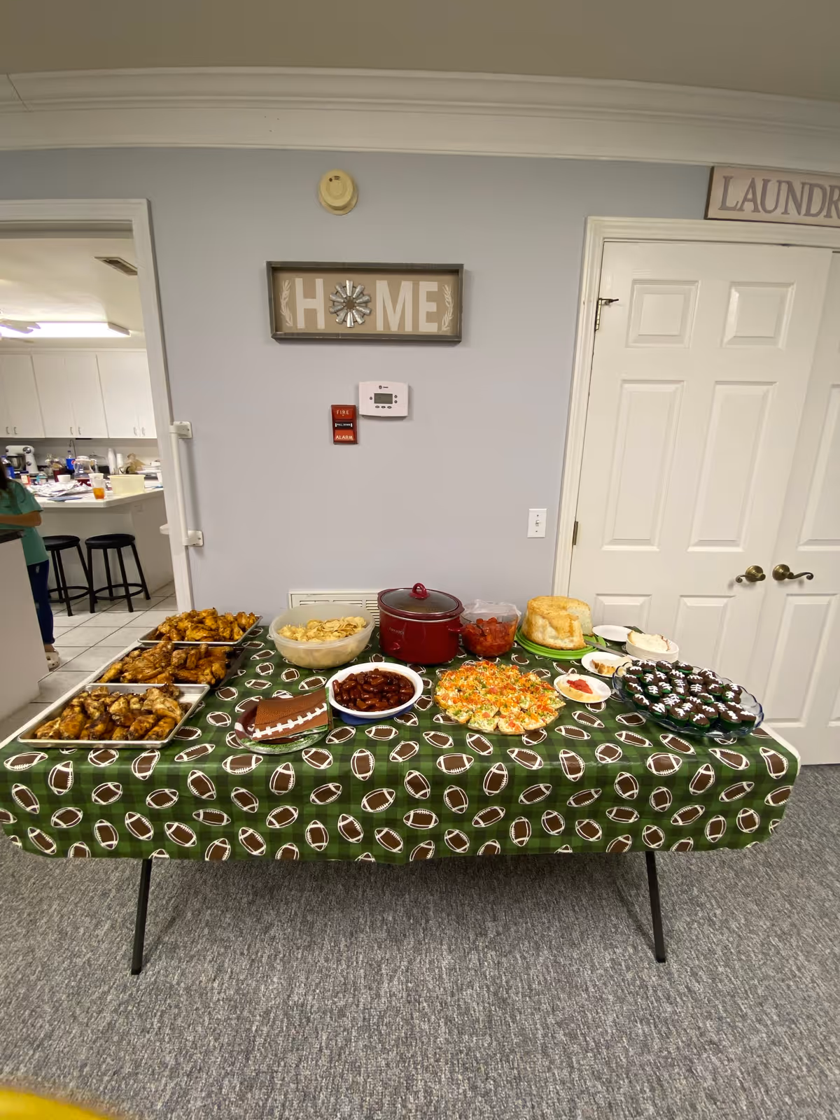 A table covered with a green football-themed tablecloth displaying a variety of food dishes including chicken wings, chips, chili, a layered salad, cupcakes decorated like footballs, and a cake. The table is set against a light gray wall with a 'HOME' sign, a fire alarm, and a thermostat. To the right, there is a white double door with a 'LAUNDRY' sign above it, and to the left, a doorway leads to a kitchen area with stools and cabinets.