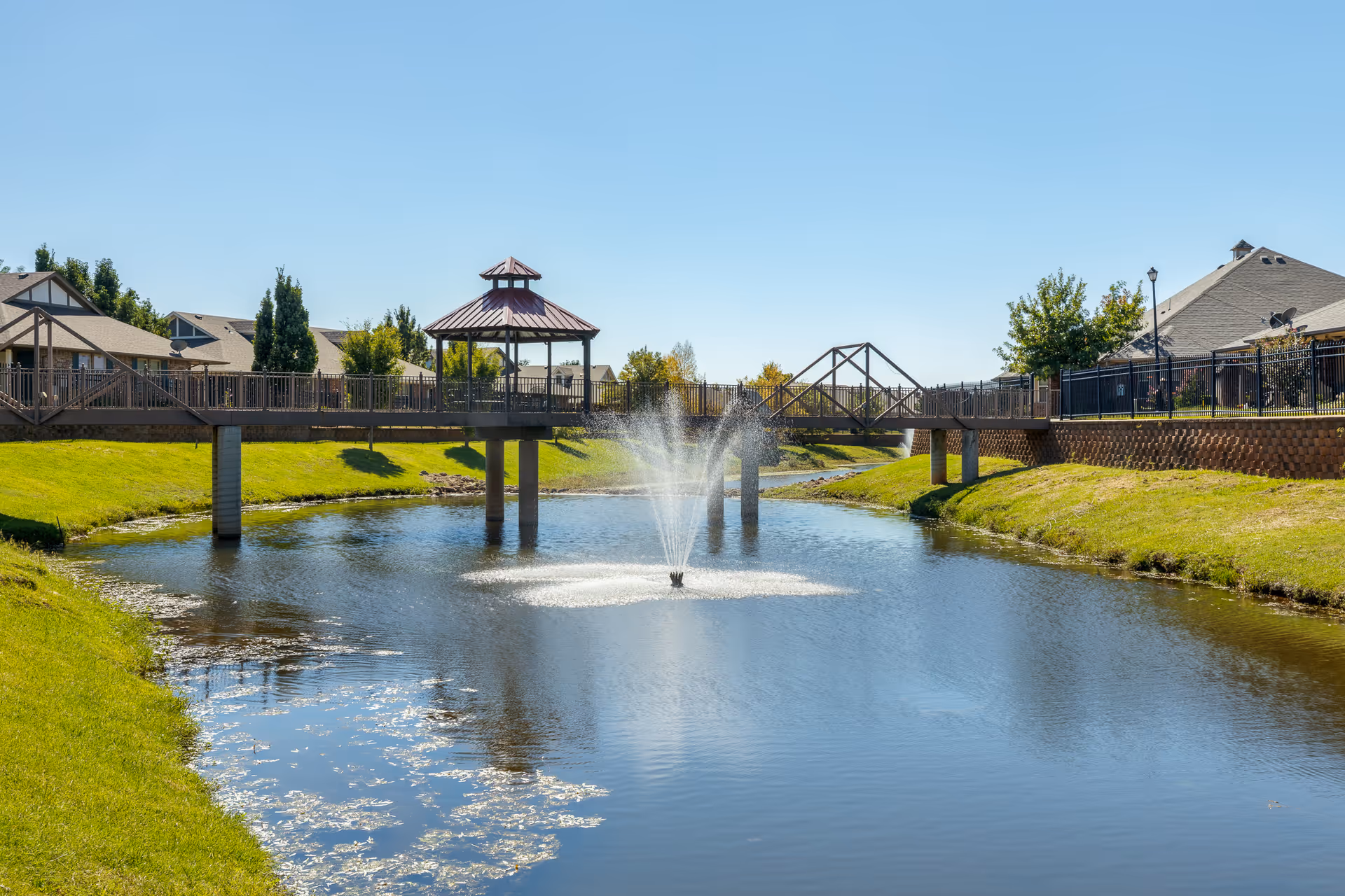 A landscaped pond with a central fountain, a wooden footbridge and gazebo, grassy banks, and nearby buildings under a clear blue sky.