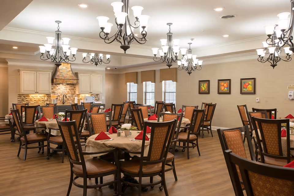 A dining room with multiple round tables covered with beige tablecloths, each set with red folded napkins and small flower arrangements. The room features wooden chairs with patterned upholstery, several ornate chandeliers hanging from the ceiling, and a kitchen area with cream-colored cabinets and a decorative range hood in the background. The walls are adorned with framed fruit artwork and windows with blinds.