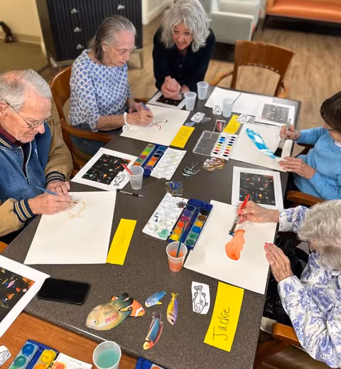 A group of elderly people sitting around a table engaged in a watercolor painting activity. Each person has a sheet of paper and paint supplies in front of them, with colorful fish images as references. A caregiver or instructor is assisting one of the participants.