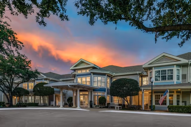 Exterior view of Wyndham Court of Plano senior living facility at sunset, showing a large two-story building with multiple windows, a covered entrance, manicured bushes, trees, and an American flag near the entrance.