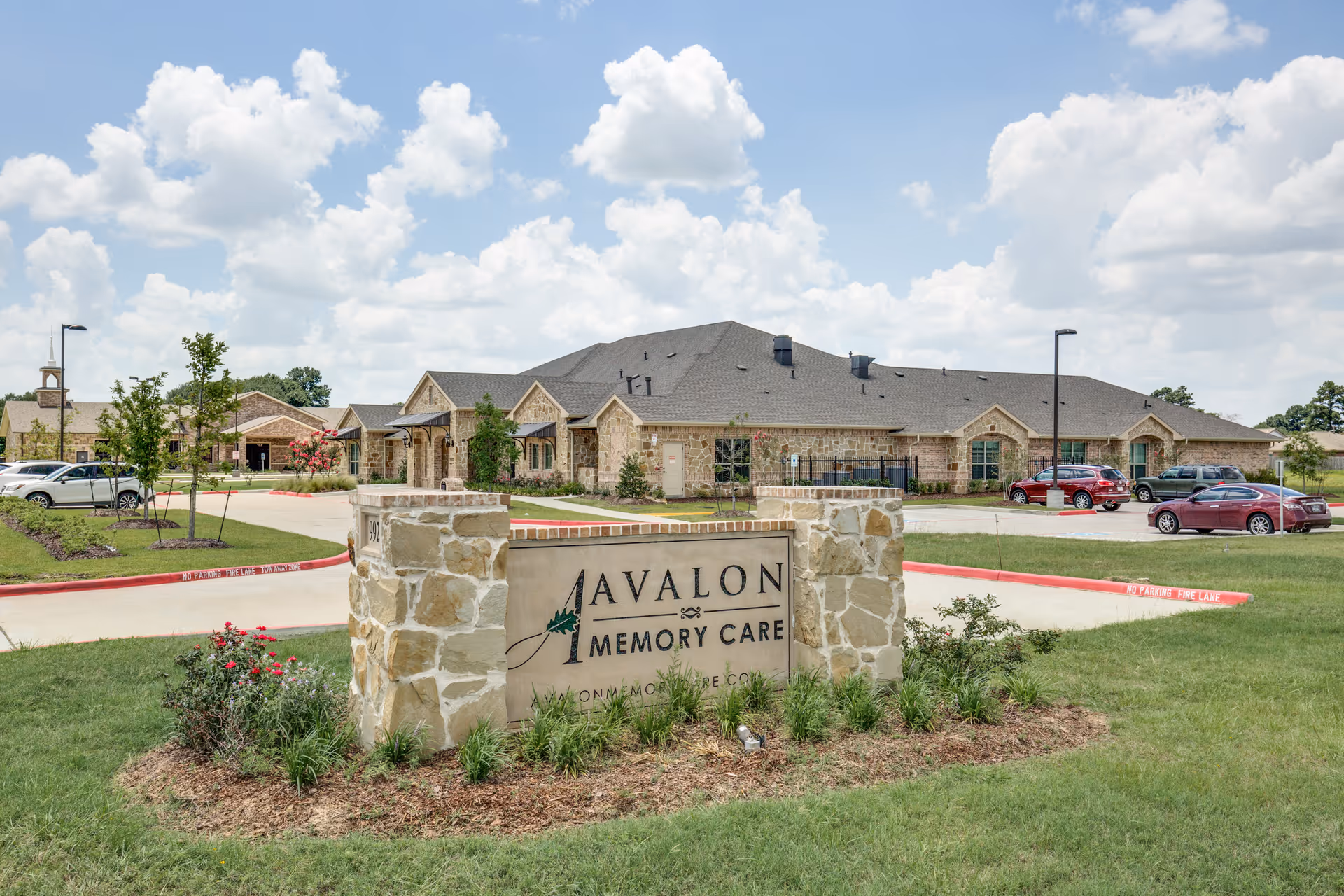 Exterior view of Avalon Memory Care facility with a stone sign in the foreground displaying the facility name. The building is a single-story structure with a stone facade, surrounded by a parking lot with several cars and landscaped greenery under a partly cloudy sky.