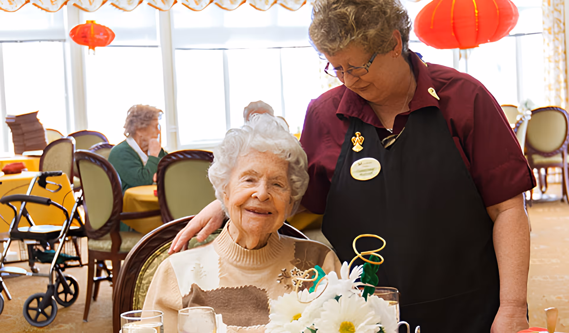 An elderly woman sitting at a dining table with a floral centerpiece, smiling, while a caregiver stands beside her with a hand on her shoulder in a warmly lit dining room decorated with red lanterns.