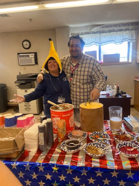 Two people standing behind a table covered with a red, white, and blue star-spangled tablecloth. One person is wearing a banana hat and a dark hoodie, smiling with arms open. The other person is wearing a plaid shirt and has an arm around the first person. On the table are various bowls of toppings, containers of ice cream, and other dessert items. The background shows an office-like room with a window, air conditioner, clock, and office equipment.