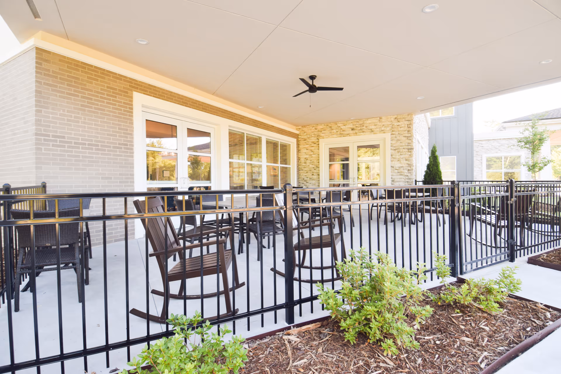 Covered outdoor patio with rocking chairs and tables behind a black metal railing.