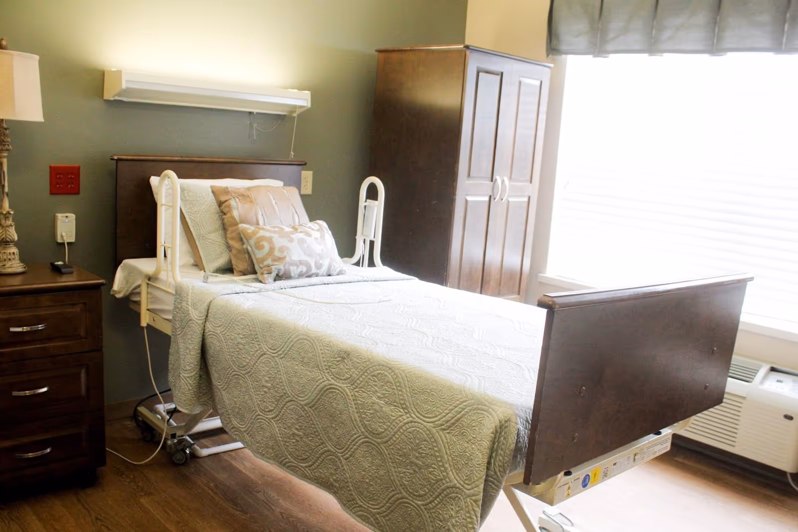 A well-lit room with a hospital-style bed covered with a light green patterned quilt and several decorative pillows. To the left of the bed is a wooden nightstand with a lamp, and behind the bed is a wall-mounted light fixture. On the right side of the room, there is a wooden wardrobe and a large window with blinds and a valance. The floor is wooden, and there is an air conditioning unit below the window.