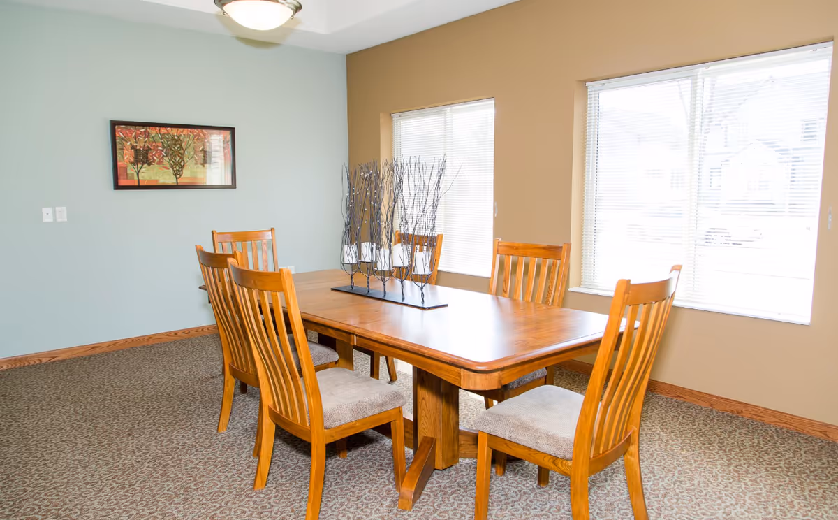 A dining room with a wooden table and six matching wooden chairs with cushioned seats. The table has a decorative centerpiece with tall, thin branches holding white candles. The room has two large windows with blinds, beige and light blue walls, a patterned carpet, and a framed artwork on the wall.