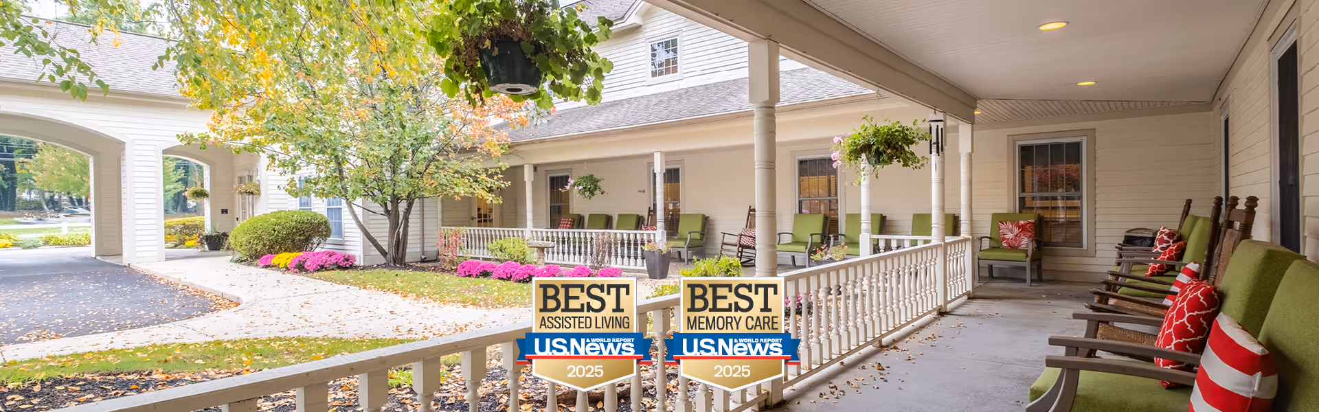Covered outdoor patio area at Benchmark at Billerica Crossings with green cushioned rocking chairs and hanging flower baskets. The patio overlooks a landscaped garden with pink flowers and a tree, adjacent to the building's exterior.